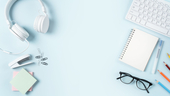 Headphones, stapler, glasses, notebook, keyboard and writing instruments against a pale blue background