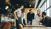 Team members gathered around a laptop in office, laughing and smiling at each other