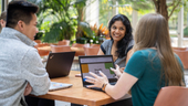 Three Workiva employees sit at outdoor table with laptops, meeting together and smiling