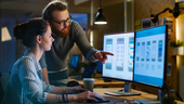 UX architect sitting a a desk having a discussion with design engineer standing next to her pointing at her computer monitor