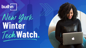 A woman sits with a laptop in front of an abstract design and the New York skyline