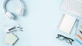 Headphones, stapler, glasses, notebook, keyboard and writing instruments against a pale blue background