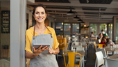 A woman in an apron leaning against the doorway of a restaurant, holding a tablet and smiling.