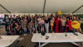 3Play team members are gathered around picnic tables under a big outdoor tent at a company event. They are wearing bright colors and have their hands up in the air.