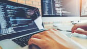 Close-up photo of software developer's hands on laptop keys with two computer screens in background showing code
