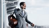 A Black man wearing a tailored suit talks on a phone at an airport while waiting for a taxi. He has a leather backpack slung over one shoul