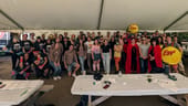 3Play team members are gathered around picnic tables under a big outdoor tent at a company event. They are wearing bright colors and have their hands up in the air.