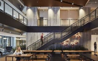 A contemporary office space with an exposed staircase. Employees work at tables and talk around the space.