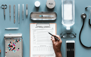a doctor holds a pen over a clipboard. Various medical supplies are organized neatly on the table around the clipboard including a thermometer, an IV bag and a stethoscope.