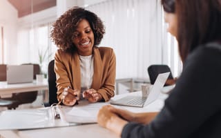Woman sits at a desk in front of another woman, explaining something on a laptop that's between them