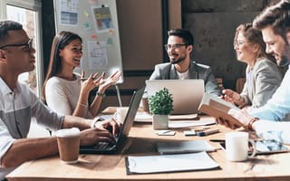 Image of five coworkers around a table smiling, talking, and typing on laptops.