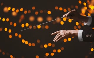 Image of a music conductor's hands using a baton with out of focus lights scattered across the background.