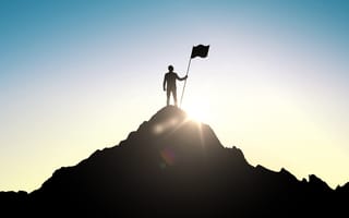 A person stands victoriously on top of a mountain holding a flag in the sunlight..