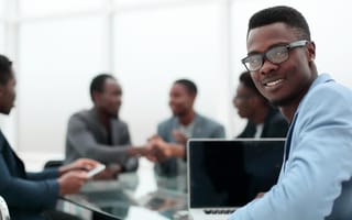 A Black employee sits at a table with colleagues and is turning to give the viewer a smile
