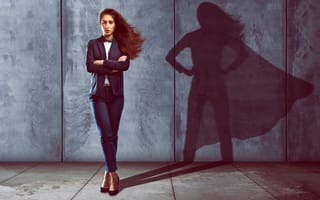 A businesswoman casting a shadow wearing a superhero cape onto the cement wall behind her