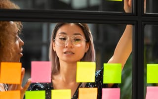 Two women at work talking about a project and looking at sticky notes on a glass window.