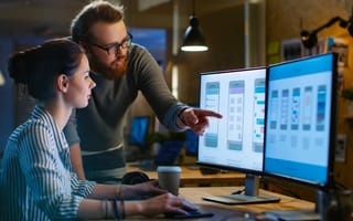UX architect sitting a a desk having a discussion with design engineer standing next to her pointing at her computer monitor