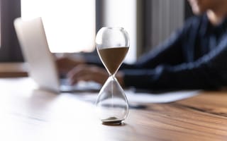 a close up of an hourglass on a wooden table with a person working on a laptop in the background