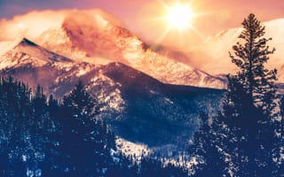 A photograph of the sun shining in the sky above a vista of the Colorado Rocky Mountains.