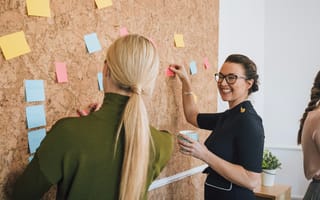 Two coworkers standing at a cork board having a discussion while posting notes