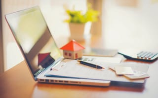 A laptop on a desk with paperwork, credit cards and a house made from paper