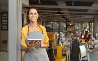 A woman in an apron leaning against the doorway of a restaurant, holding a tablet and smiling.