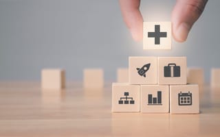 A set of blocks with images on them that represent benefits. A hand is placing a block with a plus sign on the top of the block pyramid.