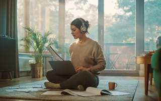 photo of person with headphones on working on laptop while sitting on the floor, with papers strewn about