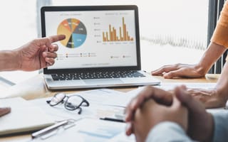 Hands from employees around a table pointing at charts and graphs on a screen.