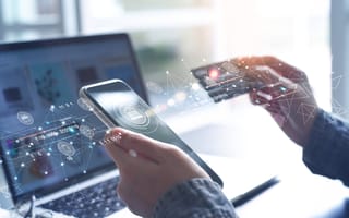 A pair of hands holding a phone and a credit card, in front of a laptop.