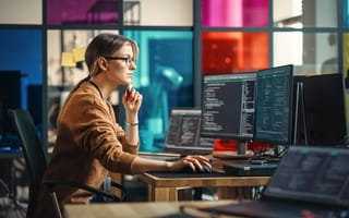 Photo of software engineer working at desk with multiple computer screens and multicolored glass wall in the background.