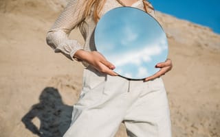 A woman in beige holds a mirror, reflecting the sky.