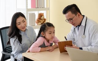 An asian woman sits in a chair with her daughter on her lap. She wears braided pigtails and a pink top. The little girl leans over a desk. An male Asian doctor is showing her something on a notepad.