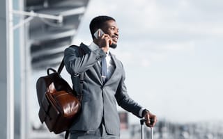 A Black man wearing a tailored suit talks on a phone at an airport while waiting for a taxi. He has a leather backpack slung over one shoulder and the handle for a rolling suitcase in the other.