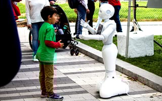 A little boy in a green shirt stands in front of a white robot. They appear to be communicating to one another.