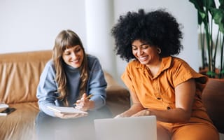 Two young professional women sitting next to each other in a casual work environment looking at a laptop together.
