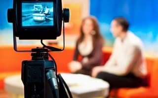 Camera records two tv hosts sitting on a sofa.