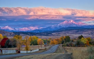 Long's Peak lights up at sunrise as a rural country road leads into the fall trees