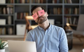 A man sits behind a laptop with sticky notes with eyes drawn on them over his own eyes.
