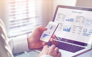 Image of a person holding a phone looking at graphs. Similar financial graphs appear on a laptop in the background.