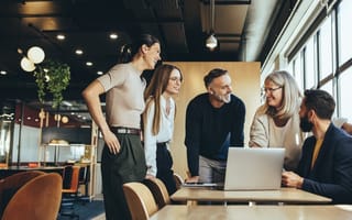 Team members gathered around a laptop in office, laughing and smiling at each other