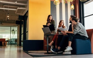 Four coworkers sit together in an office gathering area, meeting with a laptop open.