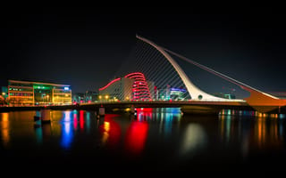 Dublin's Samuel Beckett Bridge is lit with white light at night with a brightly colored skyline in the background.