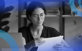 A woman at a desk with a laptop, considering a piece of paper.