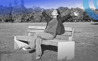 A smiling woman is sitting on a bench with her arms raised to the sky.