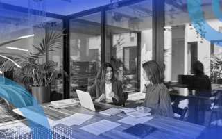 Two women talk at a table full of papers and computers