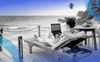 A person is sitting on a lounge chair with a laptop on a patio overlooking an ocean.