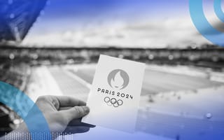 A hand is holding a card that says Paris 2024 in front of an athletic field.