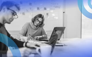 Two people are sitting at a desk working on a project. A whiteboard behind them is filled with sticky notes.