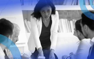 A woman professional standing to lead a meeting while her colleagues sit at the table in front of her.